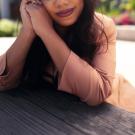 cecily is sitting at a picnic table with elbows rested on the table and hands clasped together in front of her face. she is smiling and wearing a peach suit